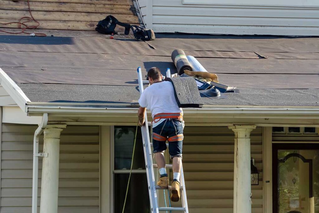 A homeowner photographs a roof leak from indoors while a worker secures a tarp outside.