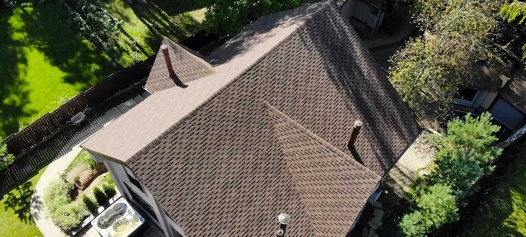 A homeowner uses a long-handled soft brush from the ground to clean moss and leaves from roof shingles.