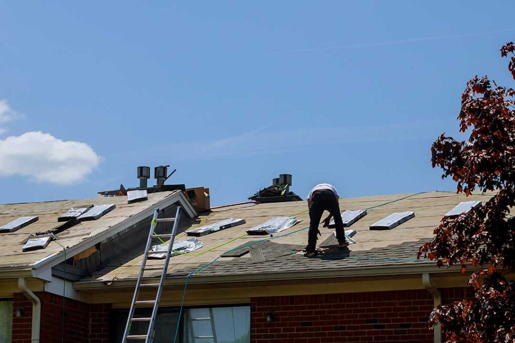 Homeowner examining a ceiling water stain beneath a roof with visible damaged shingles and wet attic insulation.
